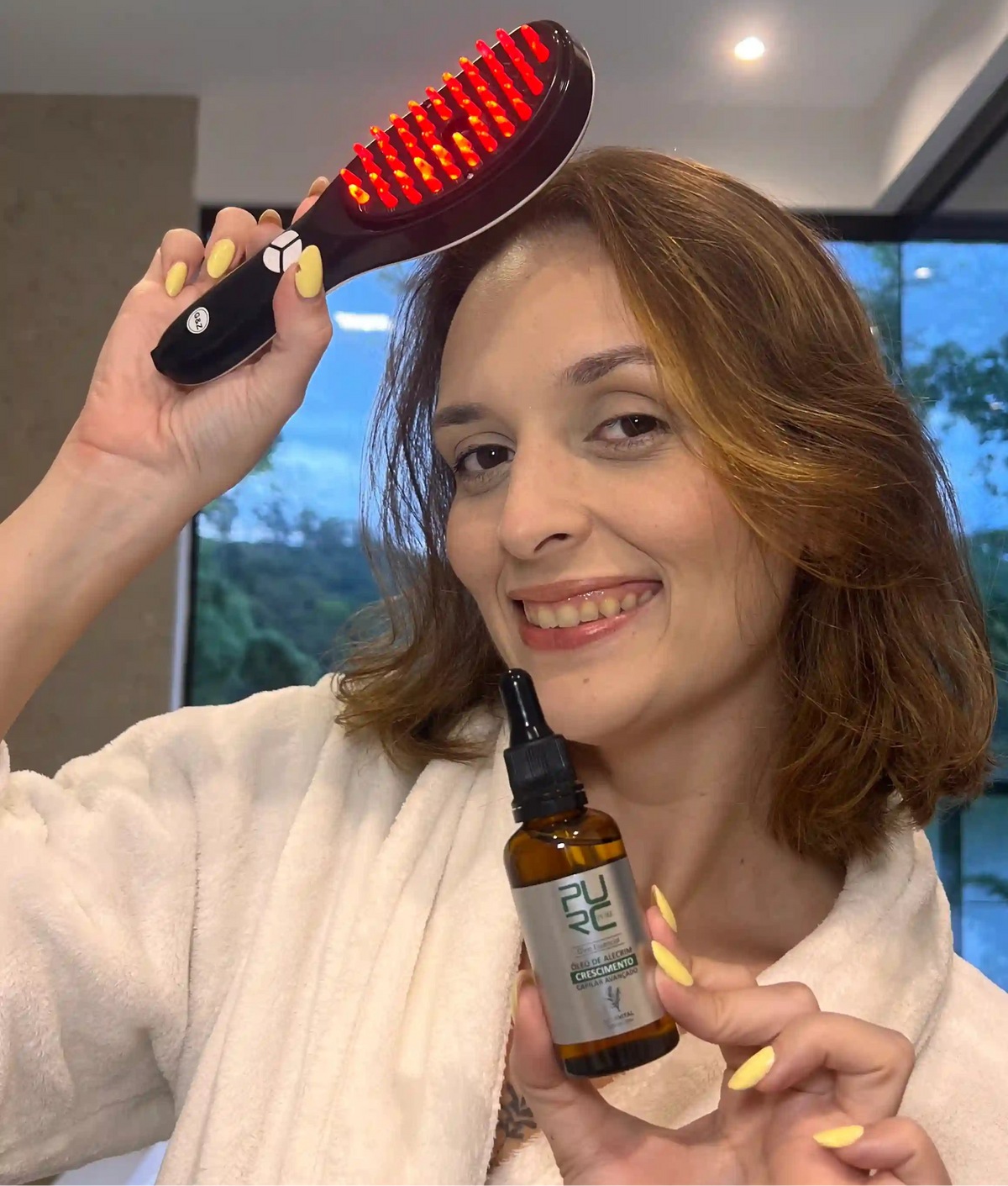 Cliente da Bela Vital segurando o kit anti quedas com a escova ligada na luz vermelha e o óleo de alecrim